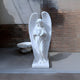 The Peaceful Dove Angel Marble Outdoor Sculpture by Giant Sculptures stands at 180cm, featuring a white marble angel holding a bird against a brick wall. The sculpture is set on a smooth gray surface with part of a stone basin visible in the background.