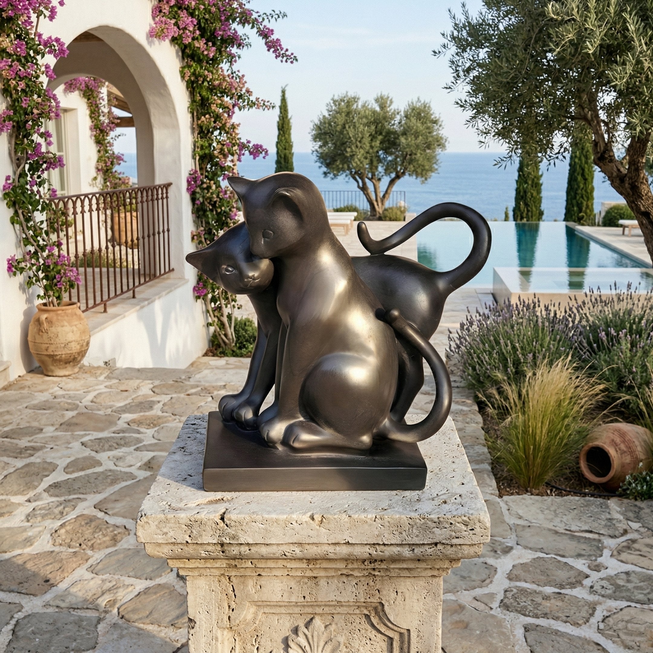 The Affectionate Cat Pair Marble Outdoor Sculpture (40cm) by Giant Sculptures stands on a stone pedestal in a Mediterranean garden with flowering vines, olive trees, garden accents, a pool, and sea views.