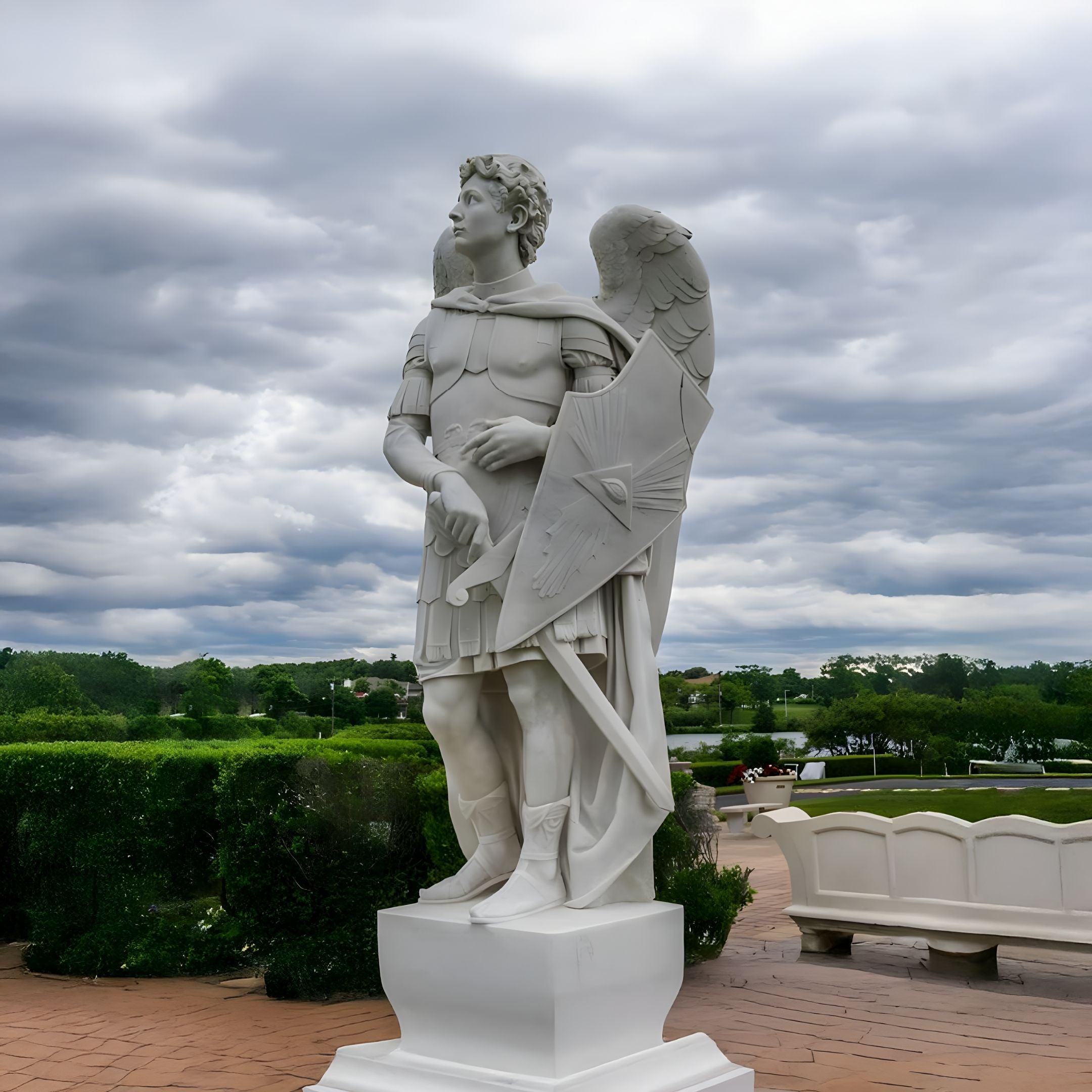 The Noble Shield Angel Marble Outdoor Sculpture - 220cm from Giant Sculptures features an angelic figure in Roman-style armor with wings unfurled, sword and shield in hand, standing on a pedestal amidst lush greenery beneath overcast skies.