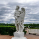 The Noble Shield Angel Marble Outdoor Sculpture - 220cm from Giant Sculptures features an angelic figure in Roman-style armor with wings unfurled, sword and shield in hand, standing on a pedestal amidst lush greenery beneath overcast skies.