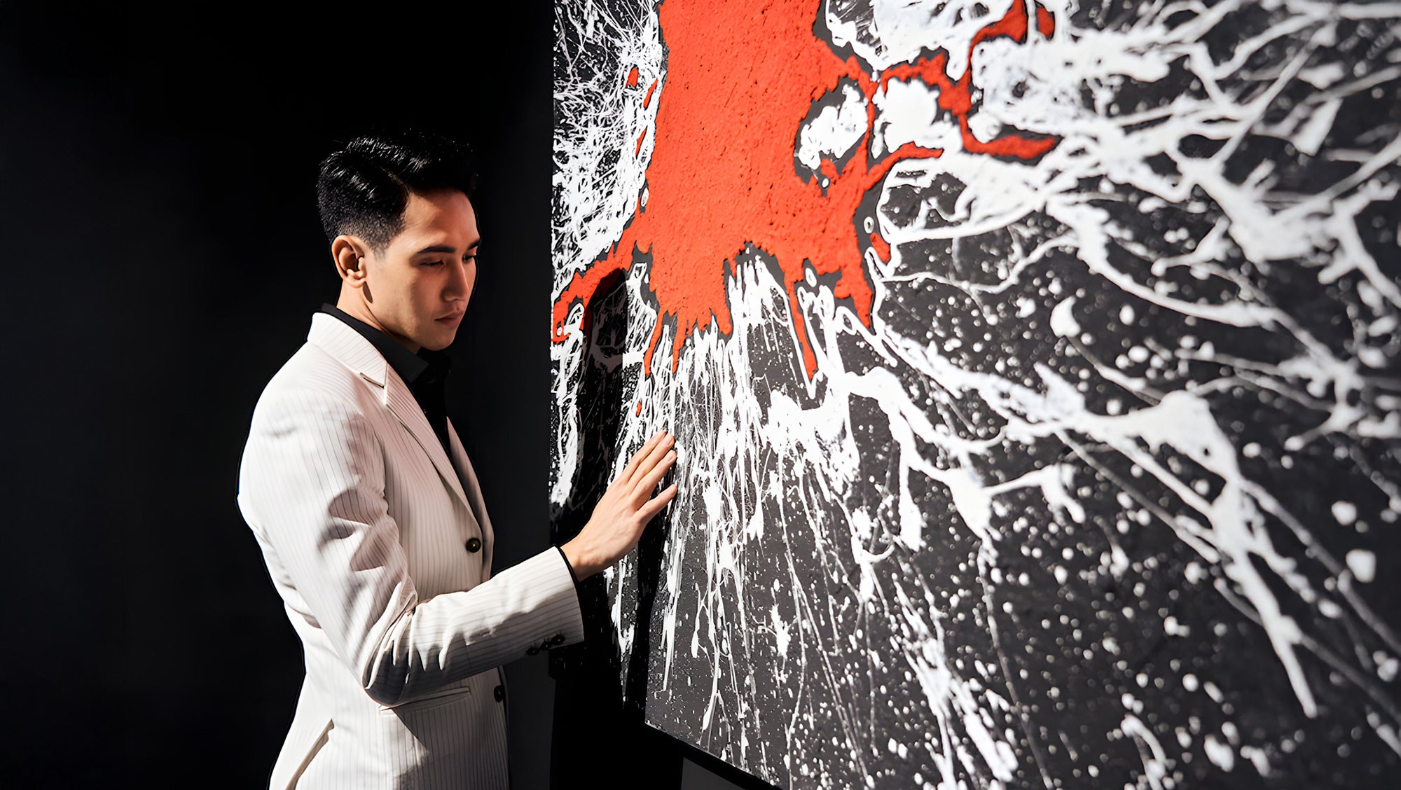 A man in a white suit stands in front of an abstract painting with red and white splashes on a black background. He touches the edge of the artwork, looking at it thoughtfully. The scene is dimly lit, focusing on the paintings texture.