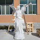 The Triumphant Trumpet Angel Marble Outdoor Sculpture by Giant Sculptures, standing at 190cm, features serene robes and wings while basking in sunlight and casting shadows against a windowed building.