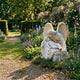 Mother And Son Angel Marble Outdoor Sculpture by Giant Sculptures, 130cm white marble angel with wings cradling a sleeping child, displayed in a lush walled garden with flowering borders.
