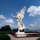 The Giant Sculptures Giant Soaring Laurel Angel Marble Outdoor Sculpture - 300cm, a large white statue, raises a laurel wreath against a partly cloudy blue sky. Situated on a landscaped platform, it is encircled by lush greenery with distant glistening water.