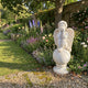 Cherub Angel Marble Outdoor Sculpture by Giant Sculptures, depicting a winged cherub seated on a sphere with hands resting on chin, placed in a walled English garden with flowering borders.