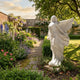 Flying Angel Marble Outdoor Sculpture by Giant Sculptures, 180cm white marble angel in mid-flight with billowing robes and spread wings, shown in a walled English garden with colorful flower borders.