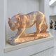 The Striding Tiger Yellow Marble Outdoor Sculpture (60cm) by Giant Sculptures stands on a stone pedestal in a modern, white-walled gallery, with abstract human-shaped statues visible in the background.