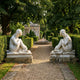 Two Seated Bathing Woman marble sculptures by Giant Sculptures displayed on stone plinths flanking a gravel garden path leading to a hedged fountain.
