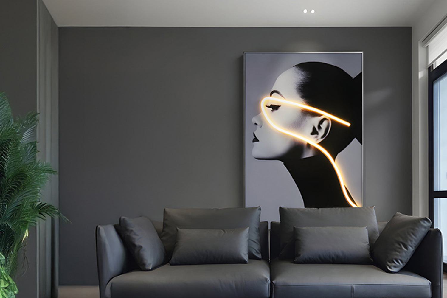 A modern living room with a dark gray sofa. On the wall hangs a black and white portrait of a womans profile with a neon light art detail accentuating her face. A green plant is visible in the corner, and a large window allows natural light in.