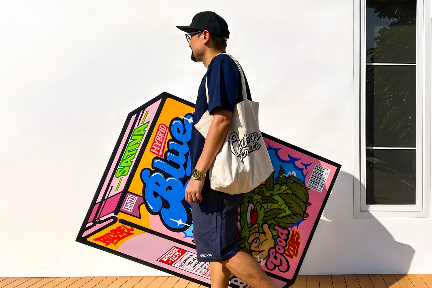 A person in a black cap, sunglasses, and navy shirt and shorts walks past a mural of a large, colorful snack box. They carry a tote bag with a graphic design. The wall is white, and theres a window with a view of leaves.