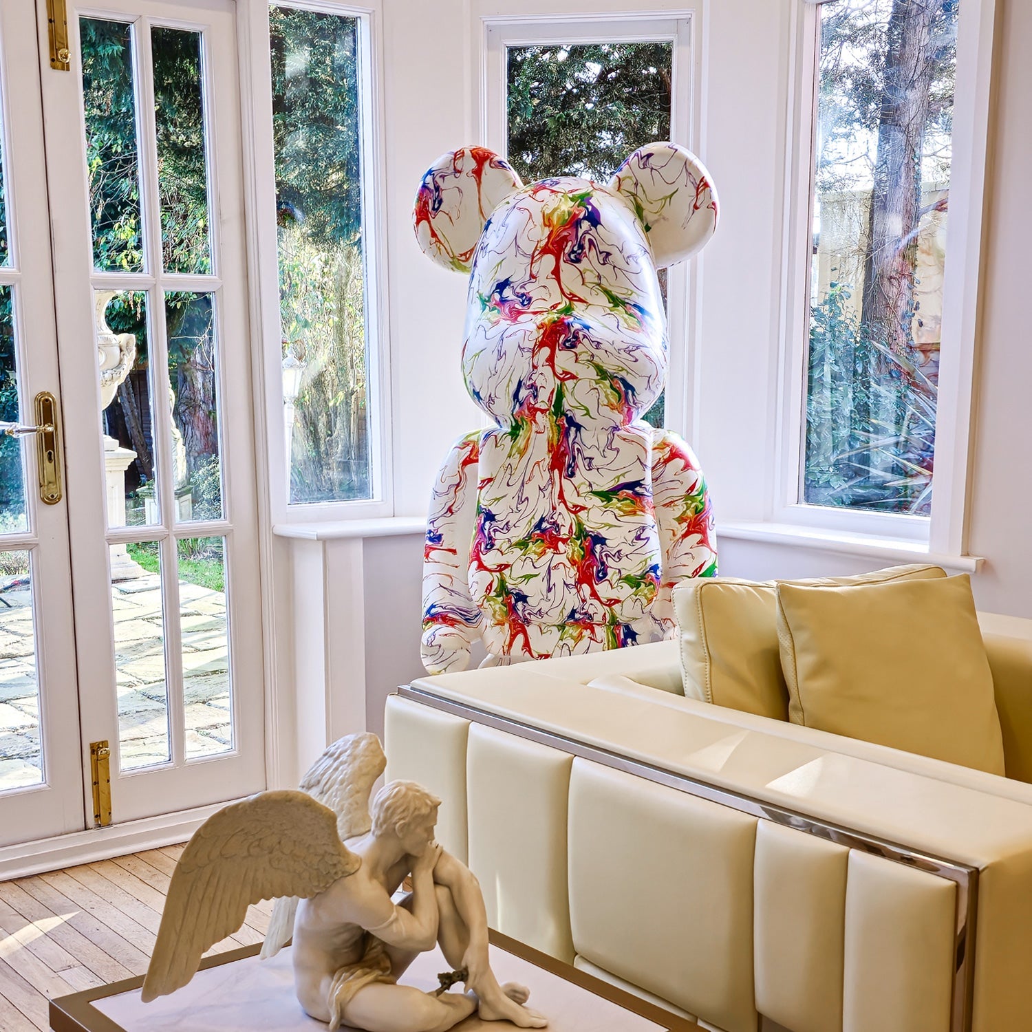 A Vogue Bear Sculpture - 165cm by Giant Sculptures with abstract designs stands in a room with large windows. In front, a small angel statue on a white coffee table complements light-colored sofas. Outside, trees sway gently in the breeze.