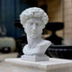 The Michelangelo's David Marble Bust Sculpture - 55cm by Giant Sculptures features a white marble bust of a curly-haired man on a square pedestal, displayed outdoors with blurred stone structures in the background.