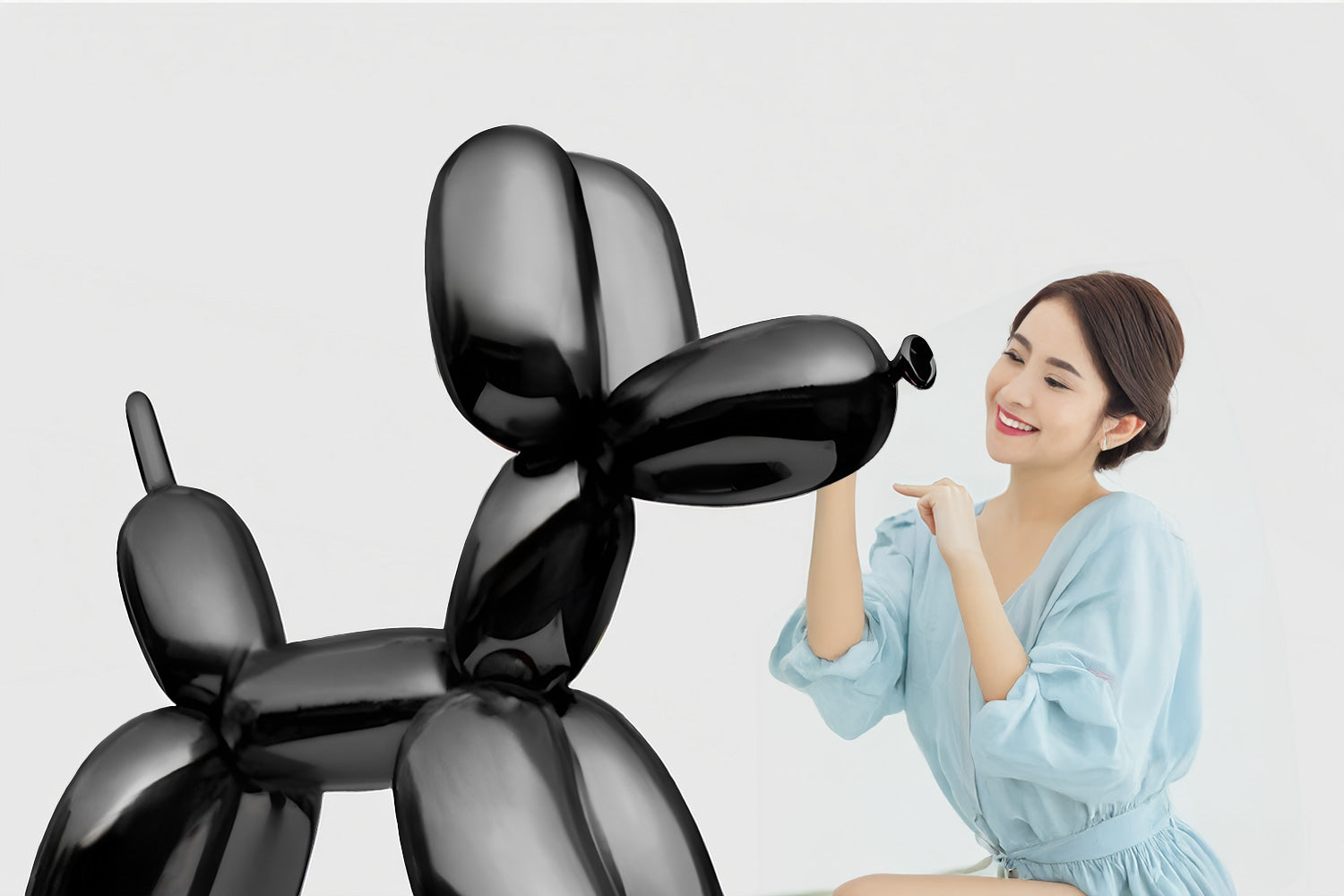 A woman in a light blue dress playfully leans toward a large black balloon sculpture resembling a dog. The background is plain white, highlighting the joyful interaction.