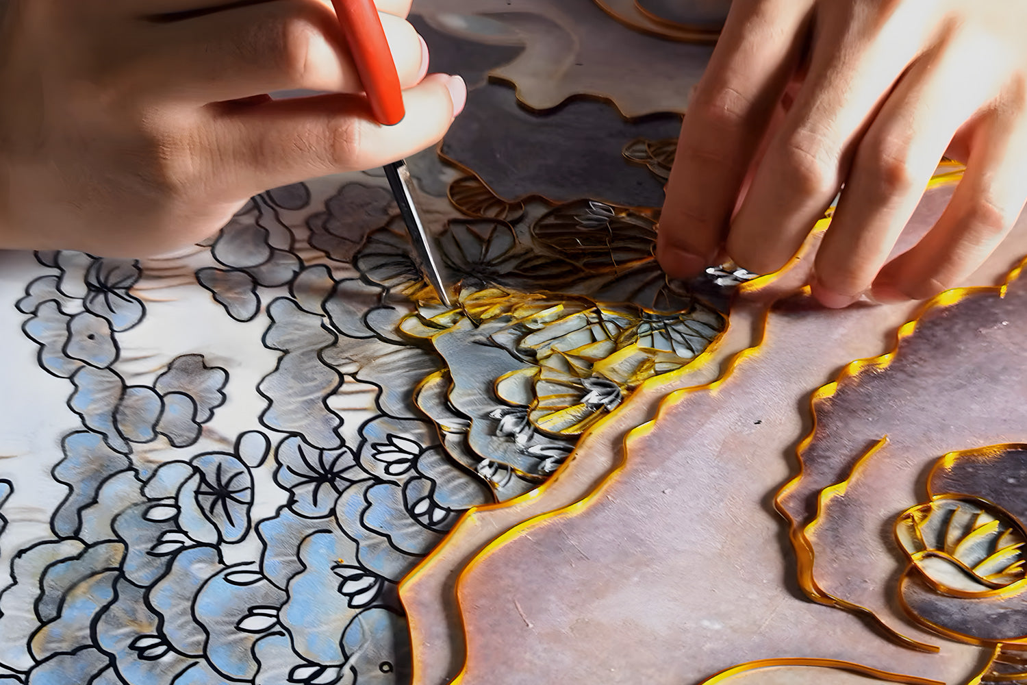 Hands using a small tool to craft an intricate design with wire and colorful materials. The artwork features floral patterns, blending shades of black, yellow, and blue on a textured surface.