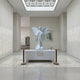 The Giant Sculptures Winged Flower Angel Marble Outdoor Sculpture (200cm) stands on a pedestal in a bright gallery with high ceilings, cream walls, and a skylight overhead; a woman's portrait hangs behind the statue.