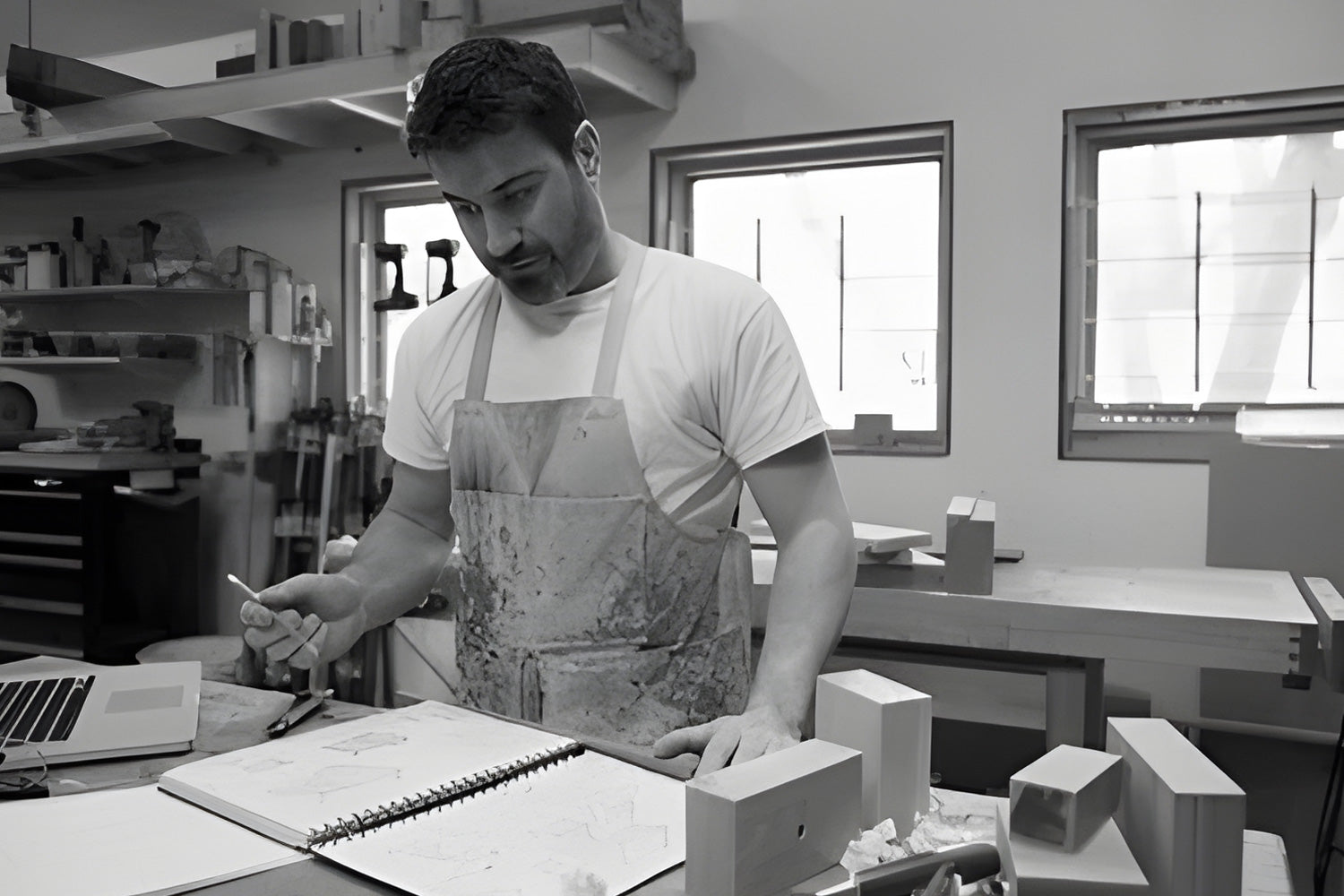 A man in a workshop, wearing an apron, stands at a table reviewing architectural sketches in a notebook. Various wooden models and a laptop are on the table, and two windows provide natural light.