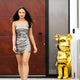 A woman in a shiny silver dress strolls outside, passing a modern building with a large wooden door, accompanied by an impressive Gilded Gold Bear Sculpture from Giant Sculptures, epitomizing contemporary art.