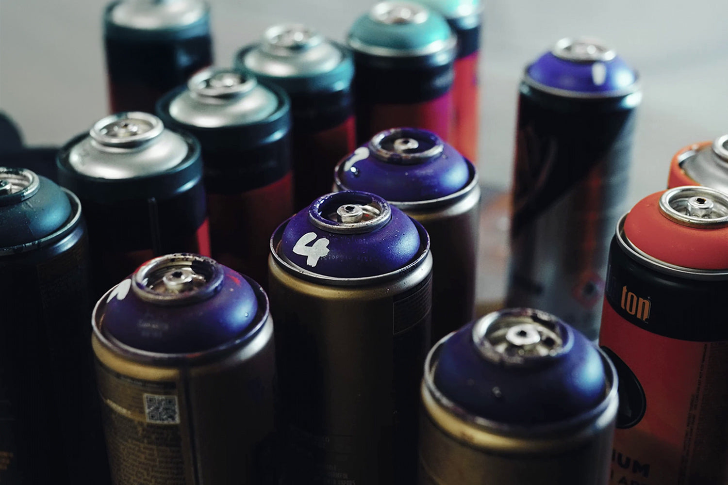 A collection of colorful spray paint cans is arranged closely together. The cans have varied colored caps, including purple, turquoise, and red. The image shows a focus on the top view of the cans, with some numbers visible on them.