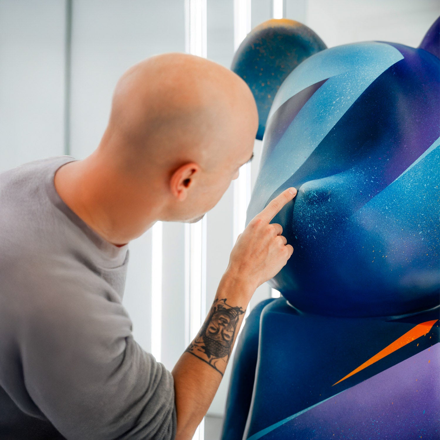 A bald person with a tattoo on their forearm examines a large, abstract blue and purple art sculpture with their finger in a brightly lit room.