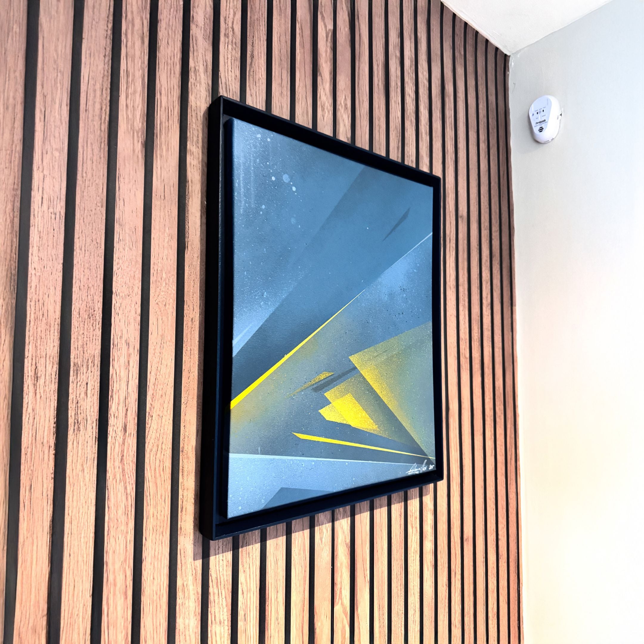 The Skull&Stone II Abstract Canvas Wall Art by Giant Sculptures, featuring blue, gray, and yellow geometric shapes, is framed and displayed on a wooden slatted wall beside a white wall with a smoke detector near the ceiling.