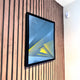 The Skull&Stone II Abstract Canvas Wall Art by Giant Sculptures, featuring blue, gray, and yellow geometric shapes, is framed and displayed on a wooden slatted wall beside a white wall with a smoke detector near the ceiling.