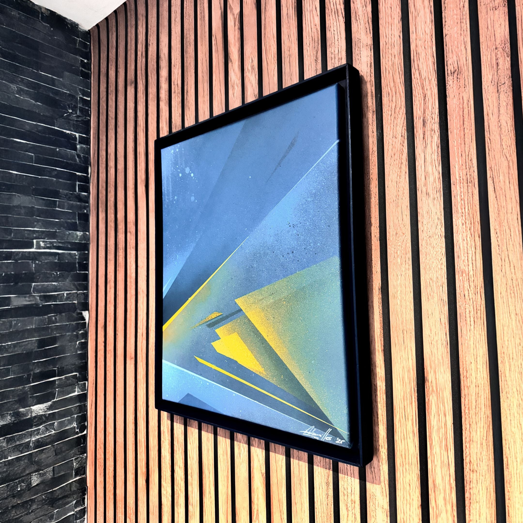 The Skull&Stone II Abstract Canvas Wall Art by Giant Sculptures, featuring sharp blue and yellow geometric shapes, is displayed framed on a wooden slat wall beside a black brick section.