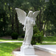 The Floral Grace Angel Marble Outdoor Sculpture by Giant Sculptures, standing at 220cm with large wings and holding flowers, is elegantly perched on a pedestal amidst lush greenery, trees, and well-maintained grass, creating a serene garden atmosphere.