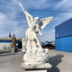 The Conquering Spear Archangel Marble Outdoor Sculpture by Giant Sculptures stands majestically at 260cm, with wings and a spear, vanquishing the figure below against blue industrial buildings and a clear sky, creating a striking contrast.