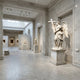 Angel Holding Cross marble sculpture by Giant Sculptures, 280cm tall, depicting a winged angel in flowing robes clasping a cross, displayed on a pedestal in a marble-clad gallery.