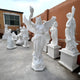 The Victory Torch Angel Marble Outdoor Sculpture by Giant Sculptures stands artistically on a pedestal, its wings spread wide. White marble statues are arranged against orange and beige walls, casting serene shadows under a cloudy sky.