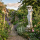 Giant Sculptures' 160cm Standing Bathing Woman marble sculpture on a weathered plinth in a cottage garden with foxgloves, roses, and a stone path to a gate.