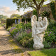 Angel With Flowers 160cm white marble outdoor sculpture by Giant Sculptures, depicting a seated winged angel with flowing robes and carved floral details, displayed in an English walled garden beside a gravel path with lavender and delphiniums.