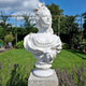 The Baroque Lady Marble Bust Sculpture - 65cm by Giant Sculptures, crafted in natural white marble with an elaborate hairstyle and draped dress, is displayed elegantly on a stone pedestal amid a lush garden of vibrant plants and flowers.