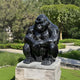 A glossy Midnight Seated Gorilla Marble Outdoor Sculpture from Giant Sculptures, featuring white veining, squats on a stone pedestal in a lush garden. Trees and grass surround it, creating a serene outdoor setting.