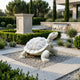 The Patterned Shell Tortoise Marble Outdoor Sculpture - 90cm by Giant Sculptures is displayed on gravel in a landscaped garden with trimmed bushes, stone paths, a waterfall, manicured trees, and a modern building nearby.
