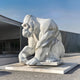 The Crouching Gorilla Marble Outdoor Sculpture by Giant Sculptures, measuring 150cm and featuring exquisite hand-carved detailing with gray veins, stands against the clear blue sky. This polished piece adds wild elegance to the modern building it guards.