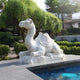 The Saddled Lying Camel Marble Outdoor Sculpture - 150cm by Giant Sculptures, crafted with intricate details, rests on a stone platform beside a pool. Surrounded by lush greenery and with a building visible under a clear sky in the background, it creates an impressive outdoor space.