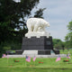 The Arctic Sentinel Bear Marble Outdoor Sculpture by Giant Sculptures stands majestically on a black pedestal, surrounded by small American flags. It contrasts strikingly against the trees and cloudy sky backdrop.
