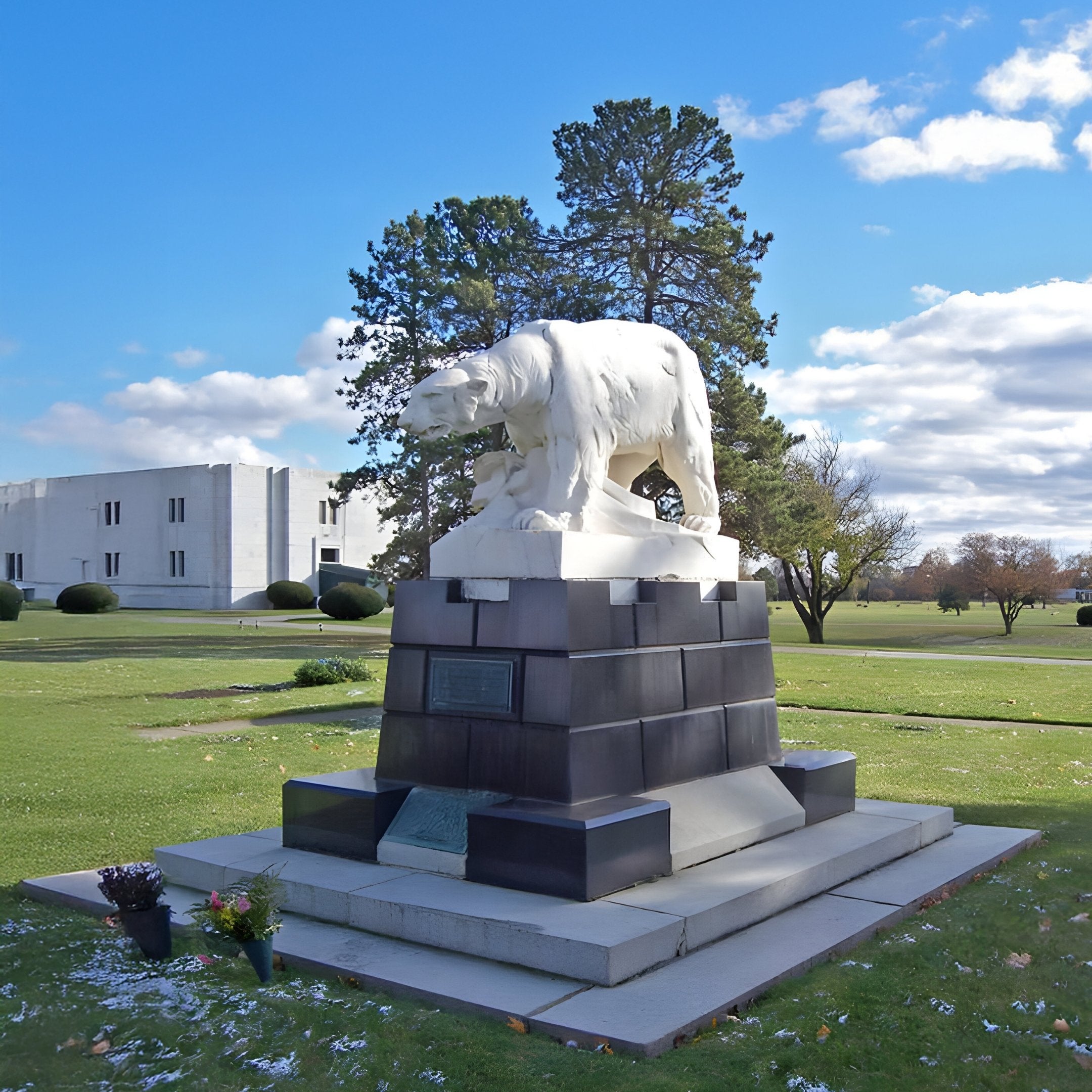 The Arctic Sentinel Bear Marble Outdoor Sculpture by Giant Sculptures stands on a tiered stone base in a grassy area, surrounded by flower arrangements. The sky is partly cloudy, and a building and trees are visible in the background, enhancing the natural beauty of this serene setting.