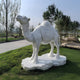 The 220cm Saddled Standing Camel Marble Outdoor Sculpture by Giant Sculptures graces a grassy area, surrounded by trees and rocks. Featuring intricate hand-carved details, the sculptures centerpiece is its saddle, set against a backdrop of clear sky.