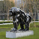 The Giant Sculptures Midnight Gorilla Marble Outdoor Sculpture - 120cm, made from Black Dali Marble, stands on a stone pedestal in a landscaped garden, reflecting light and scenery to blend seamlessly with trees and modern architecture.