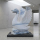 The Wings-Lifted Swan II Marble Outdoor Sculpture (120cm) by Giant Sculptures stands on a black pedestal in a modern, minimalist gallery with light grey walls and a skylight above.