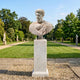 The Marcus Aurelius Marble Bust Sculpture (65cm) by Giant Sculptures stands on a pedestal in a formal garden with manicured lawns, gravel paths, and trees under a clear sky.
