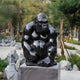 The 80cm Onyx Seated Gorilla Marble Outdoor Sculpture by Giant Sculptures, crafted from shiny black Dali marble, majestically sits on a stone pedestal in the park. Nestled among landscaped greenery and rocks, its beautifully framed by towering trees.