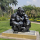 The Onyx Crouching Gorilla Marble Outdoor Sculpture by Giant Sculptures, measuring 80cm, sits majestically on a marble pedestal in a lush park. Its hand-carved geometric design is accentuated among tall palm trees and manicured hedges under a partly cloudy sky.