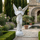 The Crowned Serenity Angel Marble Outdoor Sculpture (200cm) by Giant Sculptures stands gracefully in a formal garden, surrounded by trimmed hedges, flowers, potted trees, and tall cypress near a classical stone building.