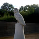 The Watchful Owl Marble Outdoor Sculpture - 40cm by Giant Sculptures is an exquisitely hand-carved piece, perched elegantly on a pedestal in a garden. It complements the wooden fence and verdant trees under a partly cloudy sky.