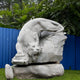 The Lunging Tiger Marble Outdoor Sculpture by Giant Sculptures stands majestically against a blue fence, hand-carved in a lifelike mid-leap pose. Its intricate details and surrounding greenery offer a perfect natural backdrop. Dimensions: 150cm.