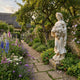 Goddess of Abundance 160cm white marble sculpture by Giant Sculptures, holding a cornucopia of grapes, set in a cottage garden with foxgloves and stone path.