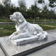 In an outdoor park, the 90cm Calm Lying Dog Marble Outdoor Sculpture by Giant Sculptures rests peacefully on a polished stone surface. The dog appears relaxed with its head raised and ears perked, while trees and pathways provide a serene backdrop.